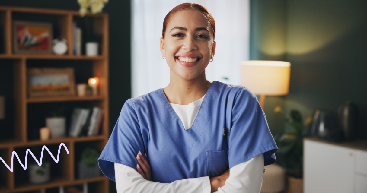 Smiling healthcare worker in blue scrubs with arms crossed, standing in a well-lit room.