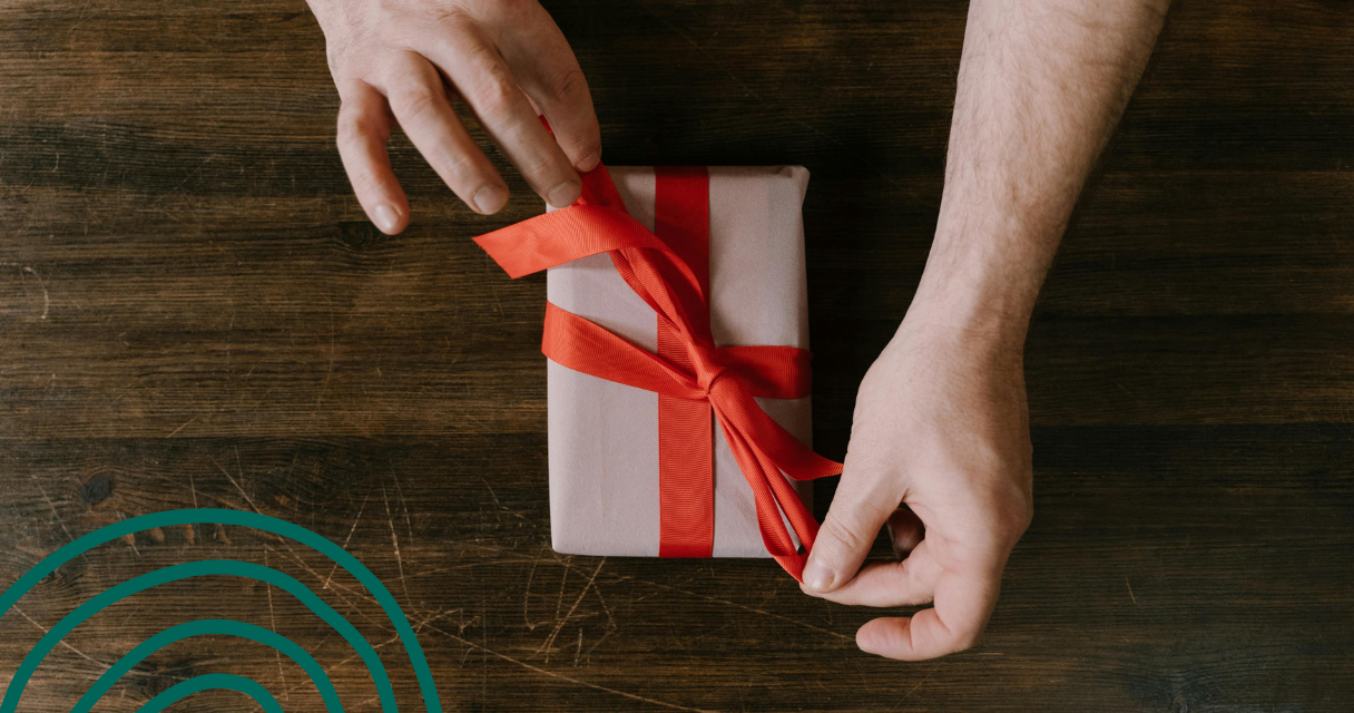 Close-up of hands tying a red ribbon on a wrapped gift, placed on a wooden table.