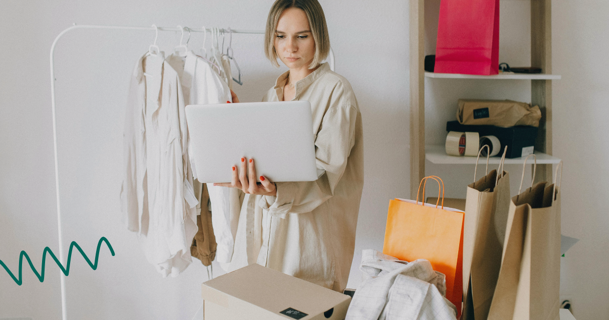 A woman standing near a clothing rack, holding a tablet and reviewing items for sale. Shopping bags and boxes are placed nearby, indicating an organized approach to selling goods to save %500 this month.