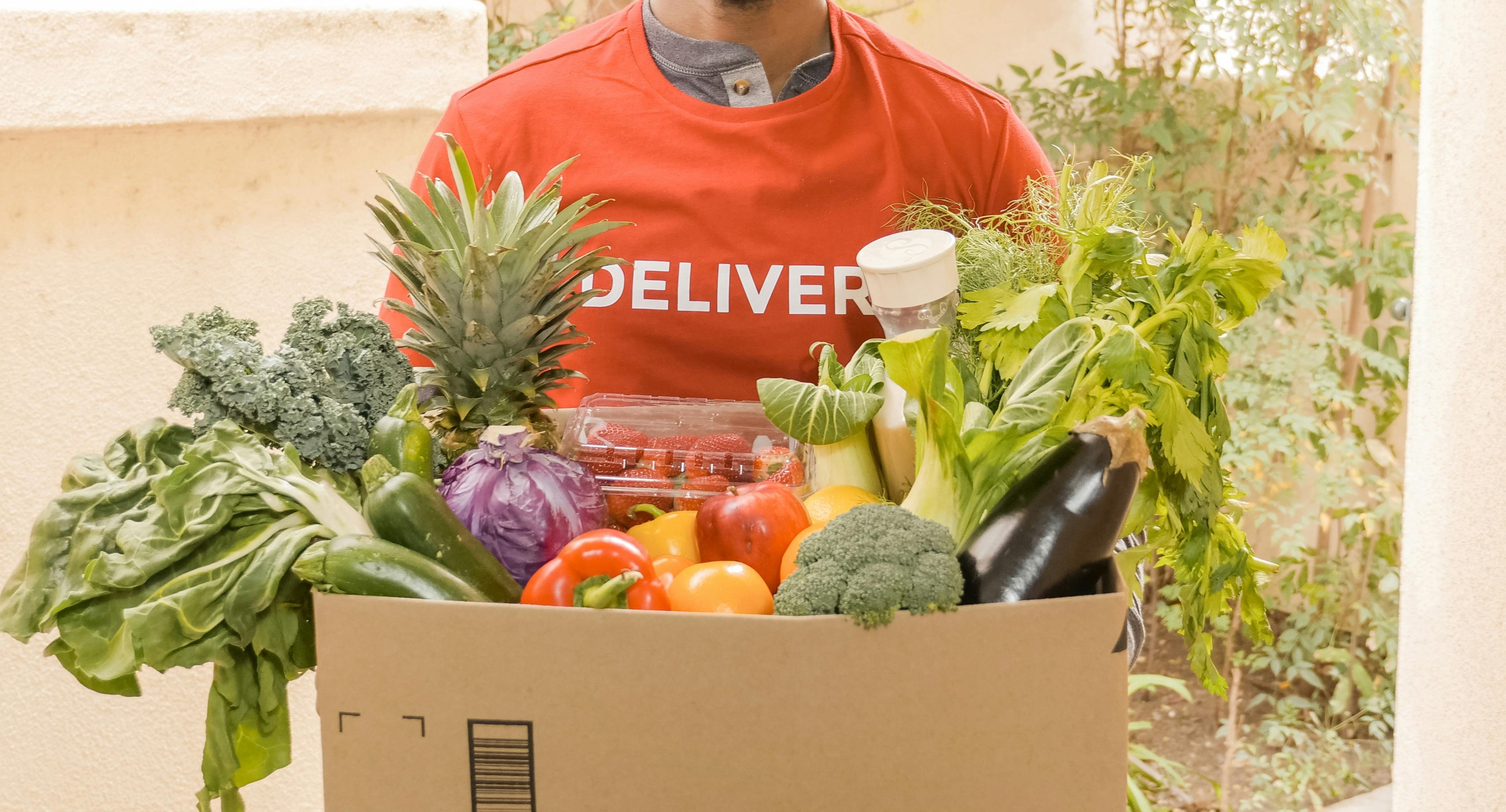 Delivery person holding a box filled with fresh vegetables and fruits, including kale, pineapple, peppers, and broccoli.