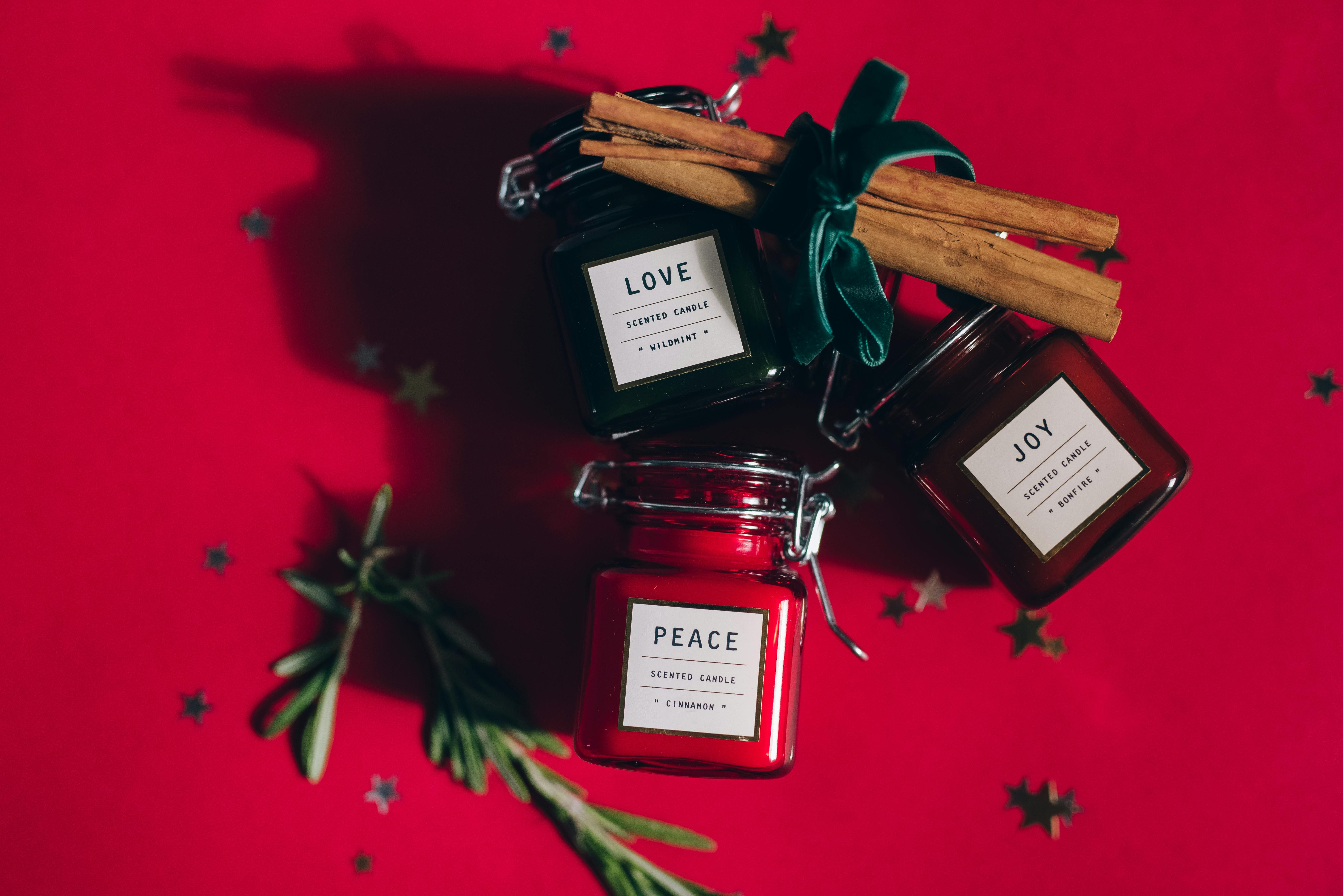 holiday tips to gift three scented candles labeled Love, Joy, and Peace on a red background with cinnamon sticks, greenery, and star-shaped decorations