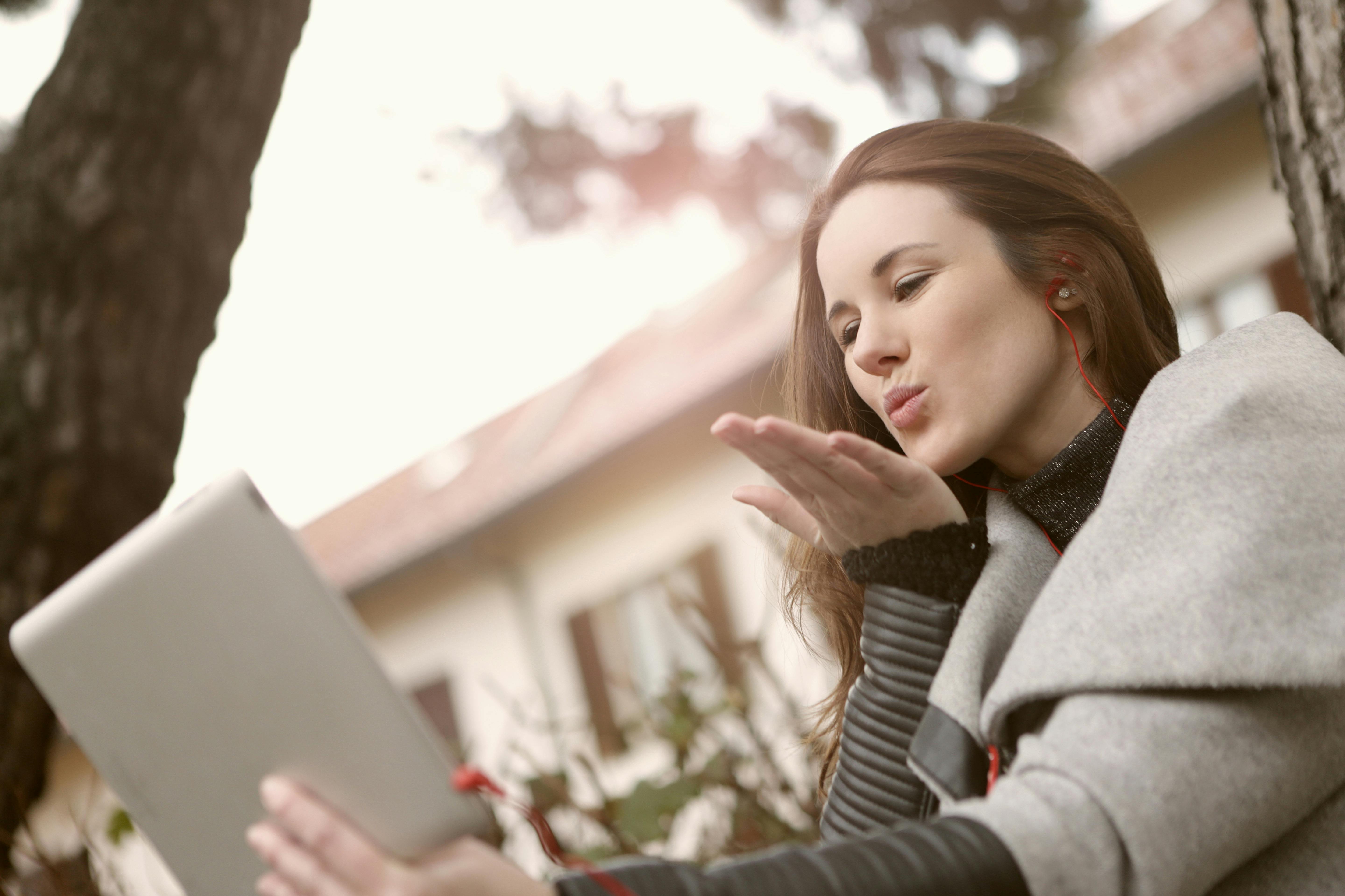 Young woman blowing a kiss during a video call on a tablet outdoors, wearing earphones and a cozy gray coat during holidays