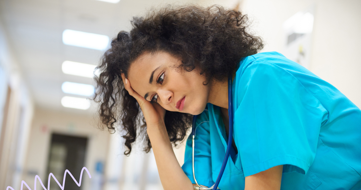 Healthcare worker in blue scrubs and stethoscope leaning forward with hand covering part of the face, blurred for privacy, in a brightly lit hospital corridor, symbolizing exhaustion and emotional toll from overwork